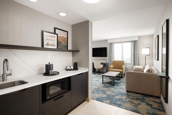 A modern hotel room with a kitchen area, a sofa, a TV, and a flat-screen TV mounted on the wall.