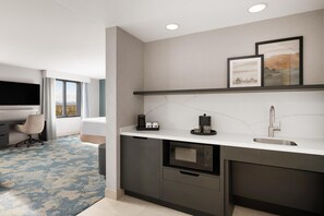 A modern hotel room with a kitchenette, a flat-screen TV, a desk, and a view of the outdoors.