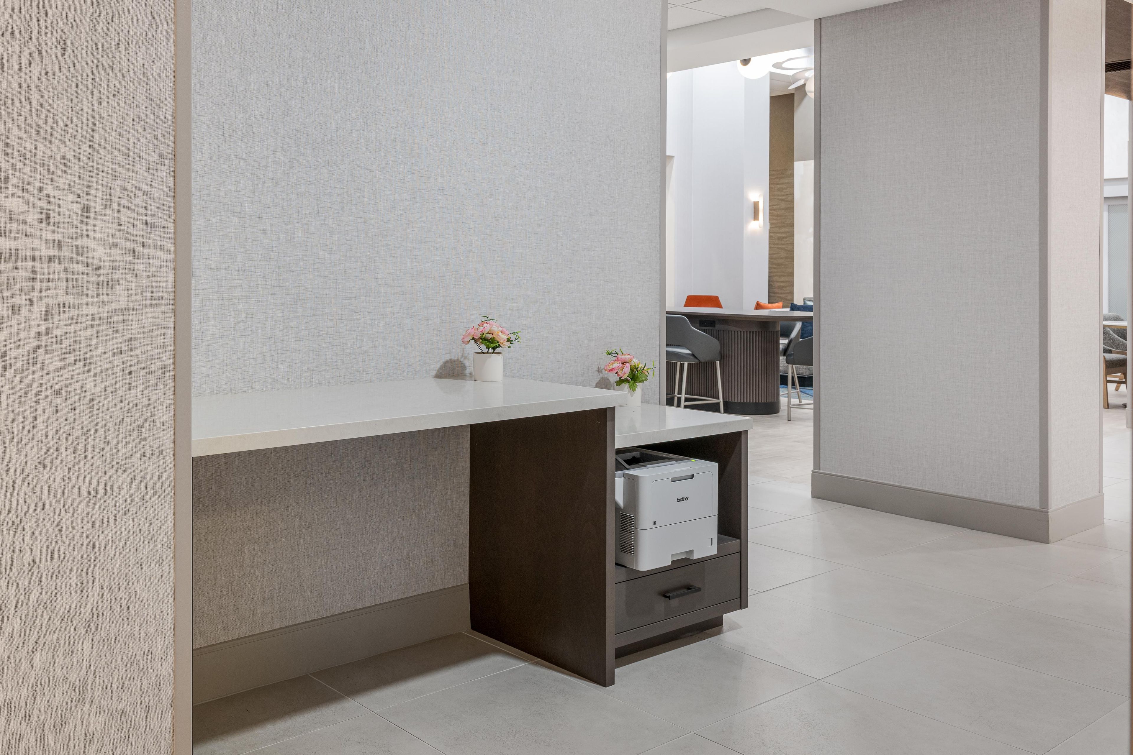 A modern office space with a white desk, a small shelf with a printer, and a vase with flowers.