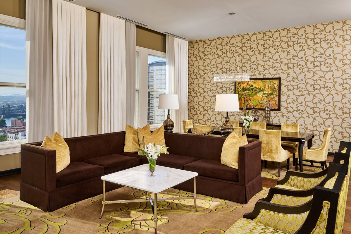 A hotel room with a brown sofa, a white coffee table, a dining table with chairs, and a large window with curtains.