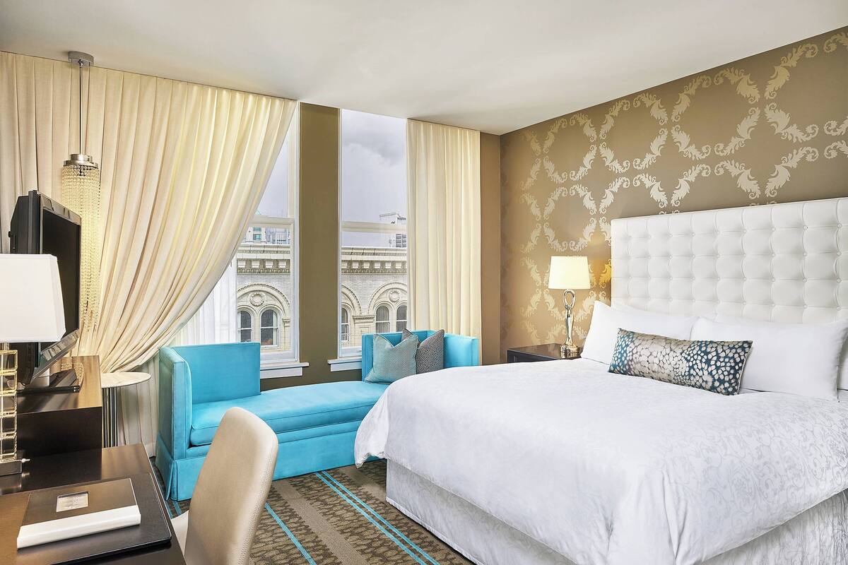 A hotel room with a large bed, a blue sofa, a desk with a laptop, and a view of a building through the window.