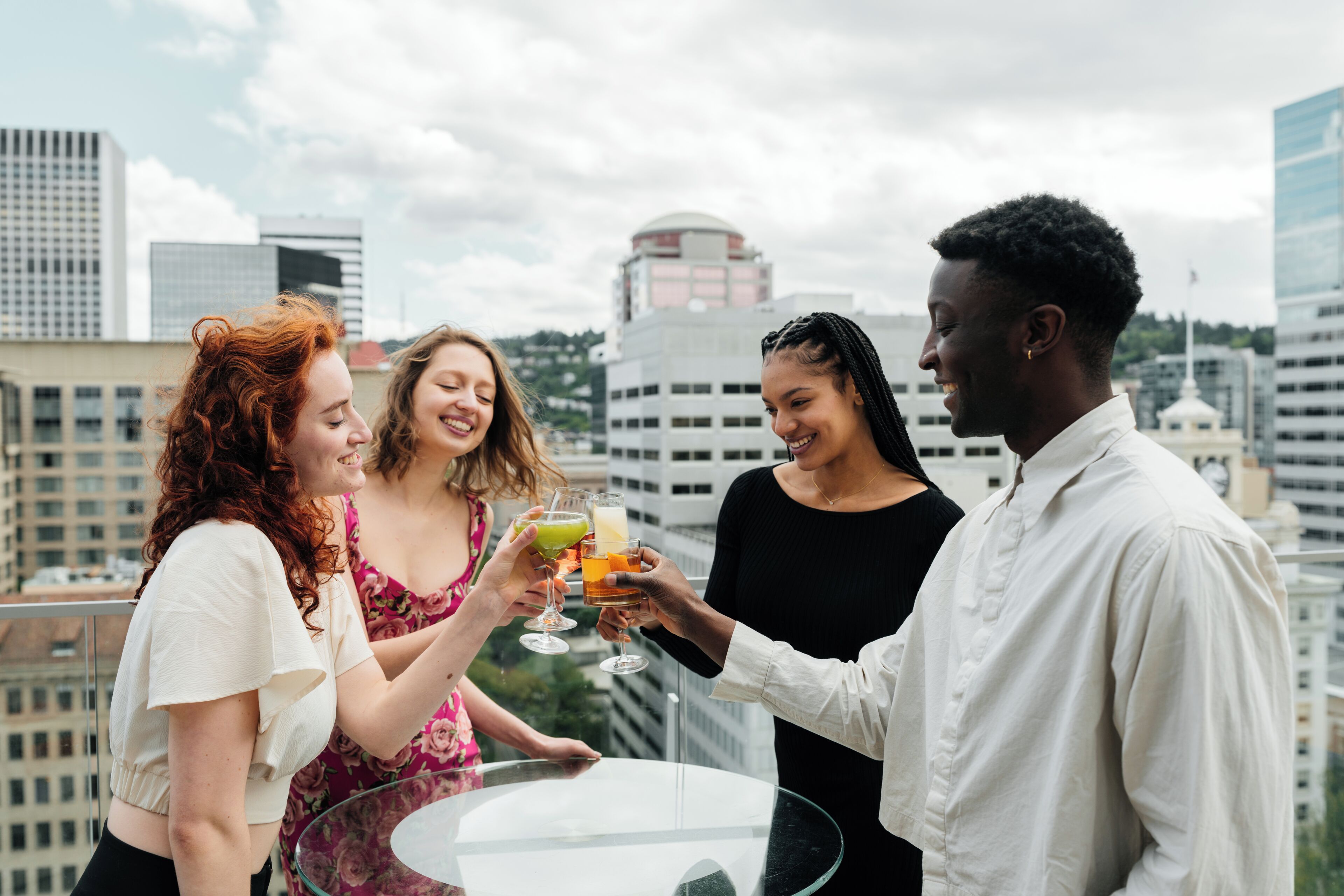A group of people toasting with drinks on a rooftop terrace with a city skyline in the background.