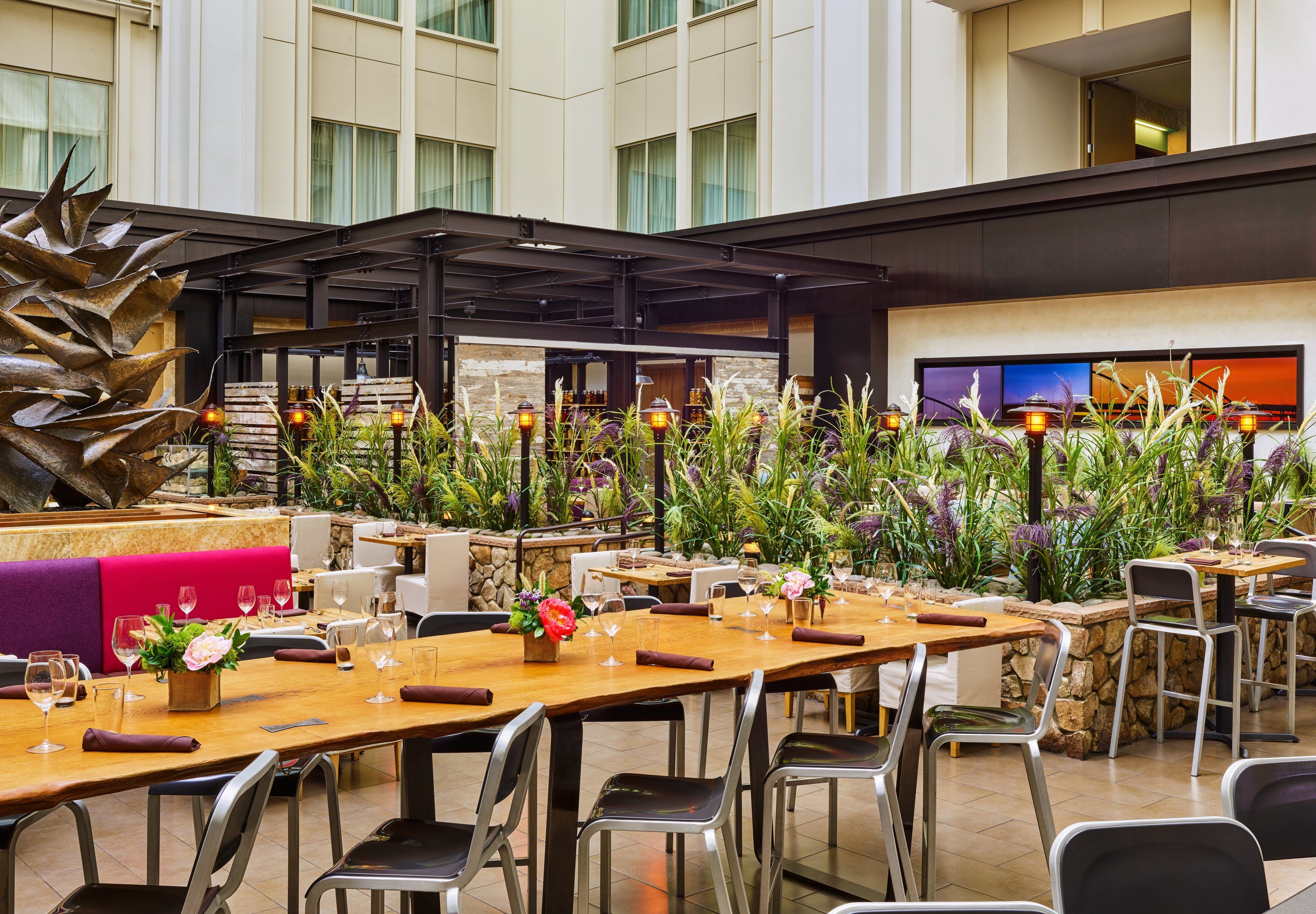 A spacious indoor dining area with multiple tables and chairs, a large decorative plant, and a stone wall with a television screen.