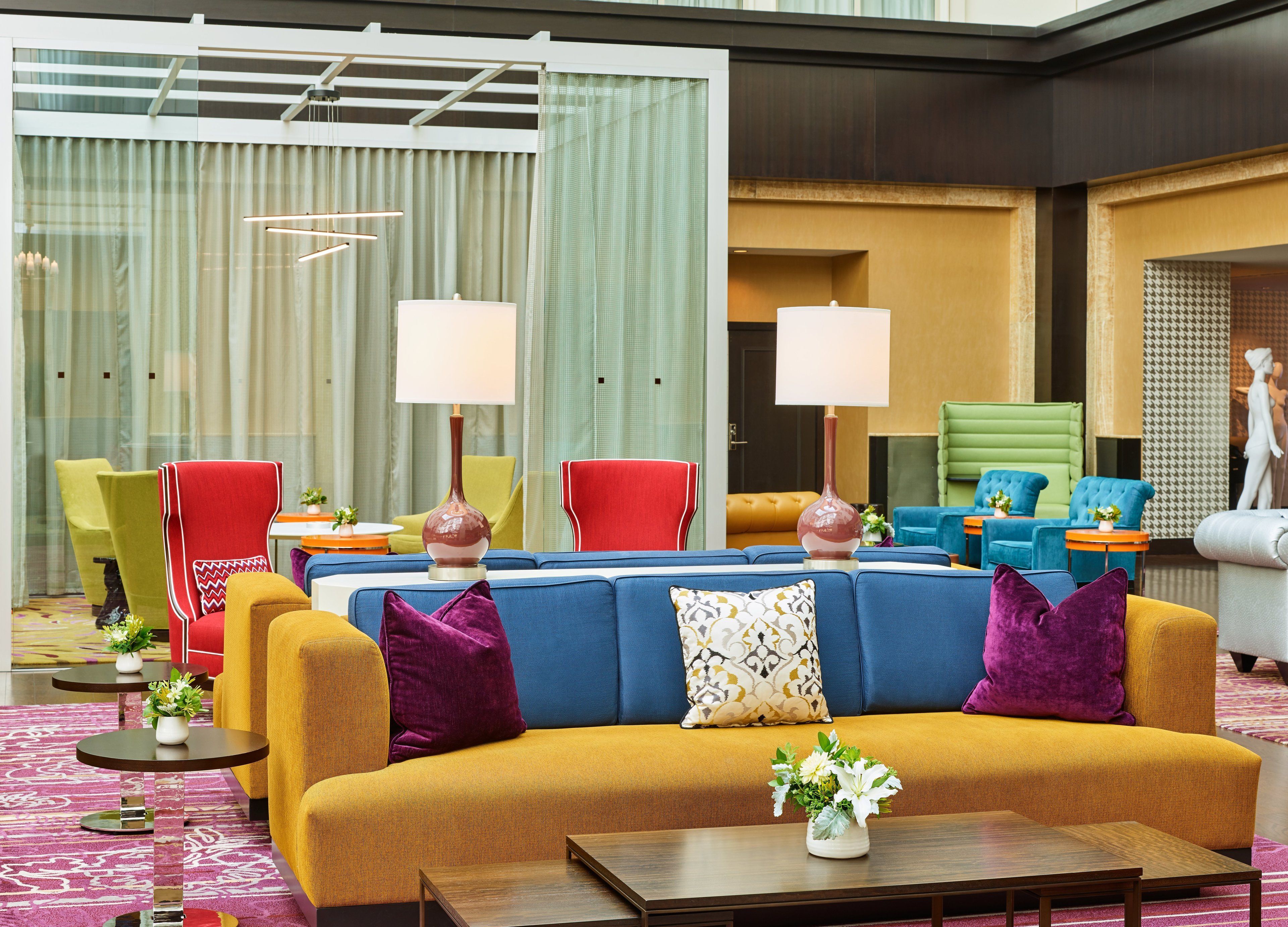 A modern hotel lobby with a blue sofa, yellow armchairs, and a red chair. There are two lamps on the sofa, a small table with a vase of flowers, and a patterned rug.