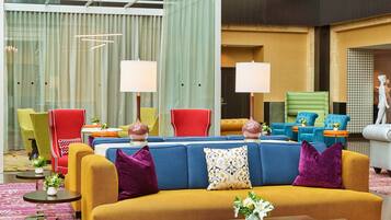 A modern hotel lobby with a blue sofa, yellow armchairs, and a red chair. There are two lamps on the sofa, a small table with a vase of flowers, and a patterned rug.