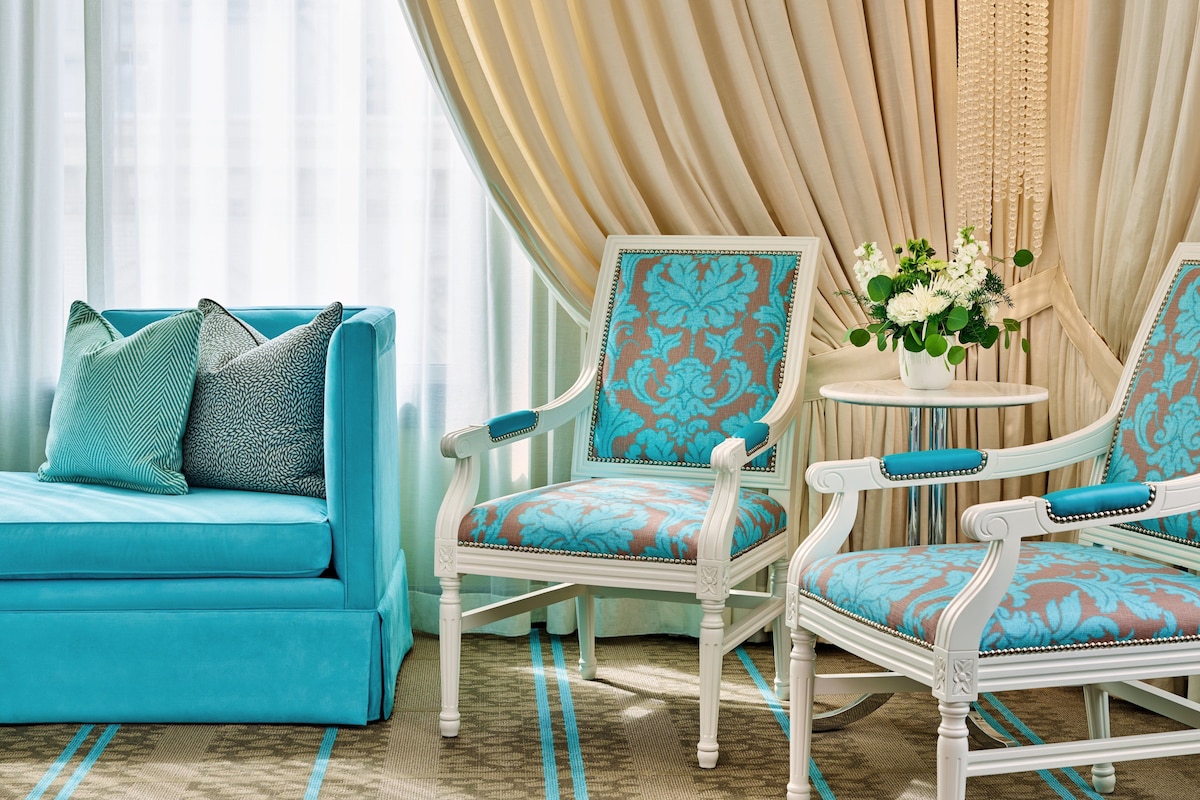 A room with a teal sofa, two patterned armchairs, a small round table with a vase of flowers, and a large window with sheer curtains.