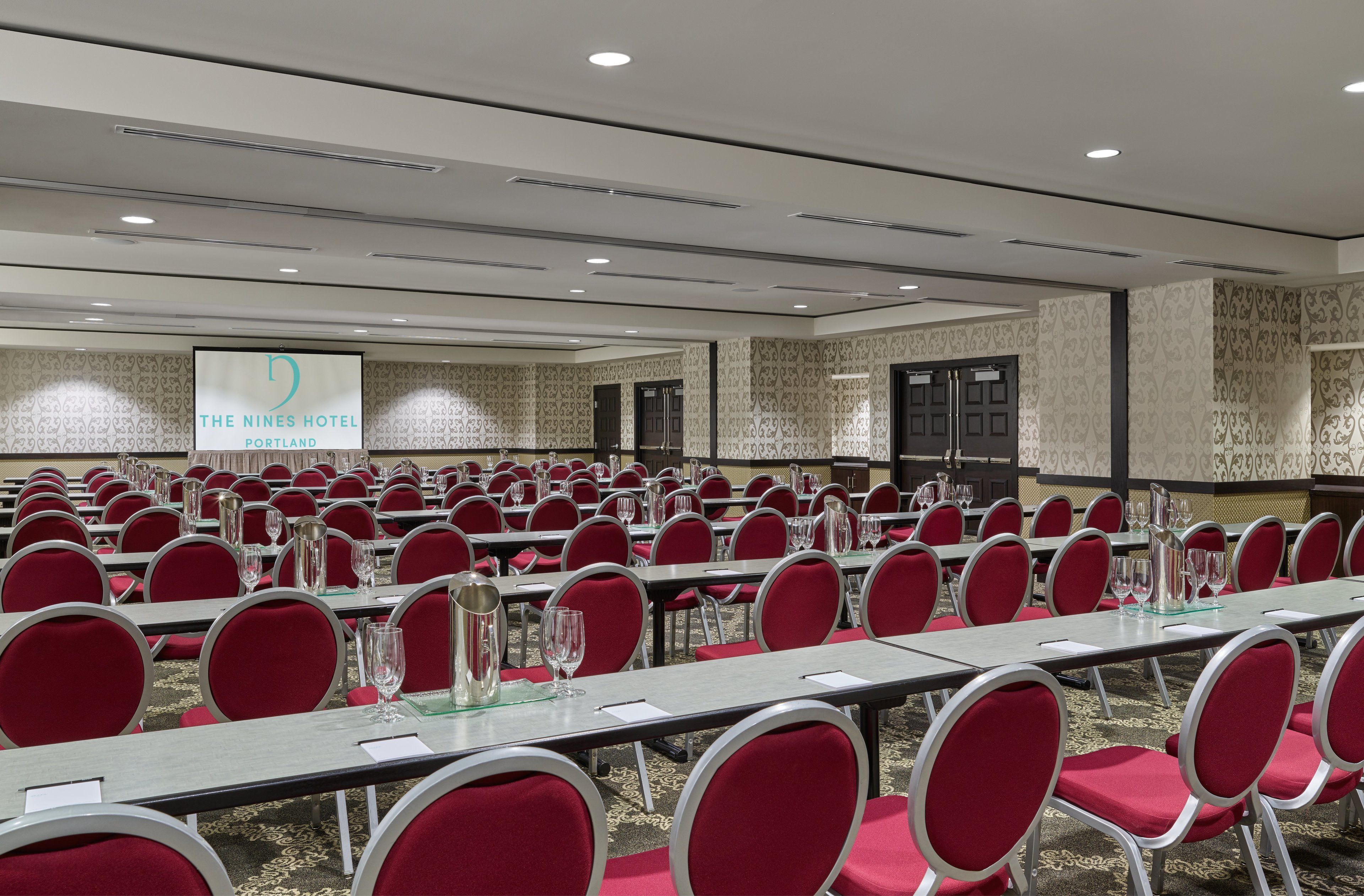 Une salle de conférence à l’hôtel The Nines, à Portland, avec des rangées de chaises et de tables rouges, un écran de projection et un papier peint à motifs.