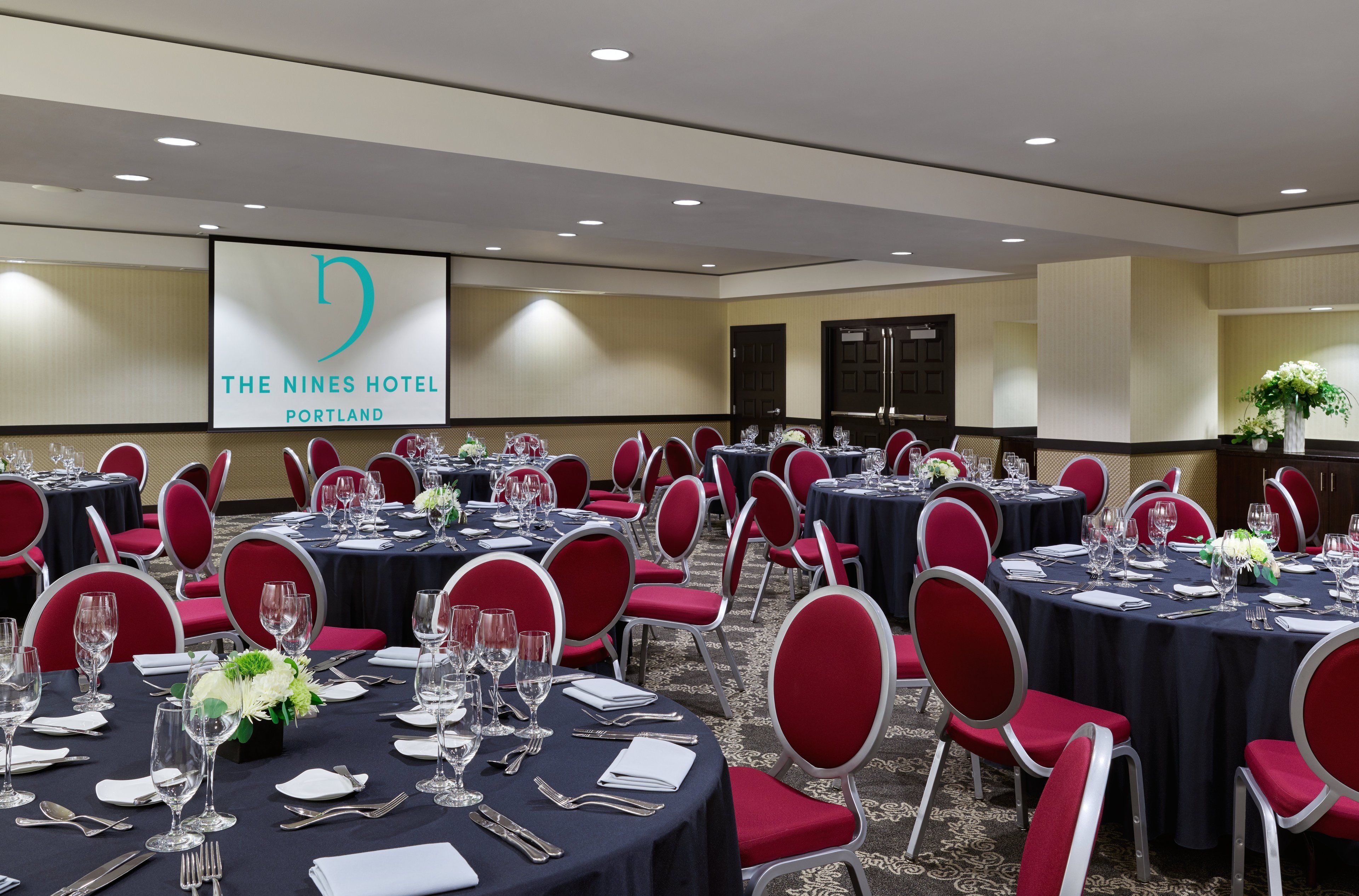 Une salle de conférence de l’hôtel The Nines, à Portland, avec plusieurs tables rondes disposées pour les repas, des chaises rouges et un écran de projection affichant le nom de l’hôtel.