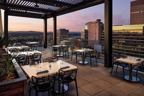 An outdoor dining area with tables and chairs, large windows, and a view of the cityscape.