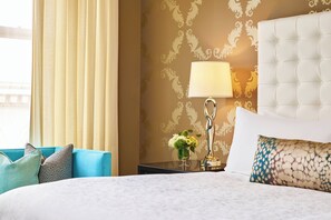 A hotel room with a large bed, a nightstand with a lamp, a vase with flowers, and a view of the outdoors.