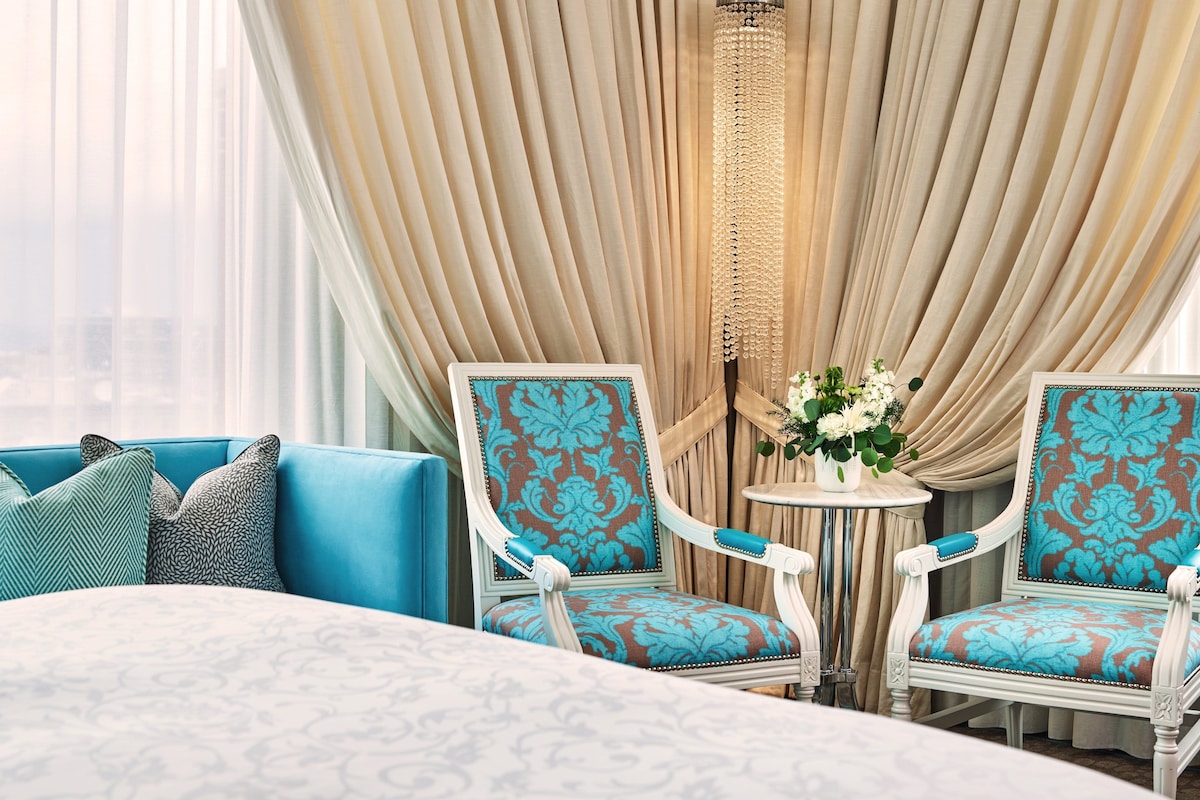 A hotel room with a large bed, two armchairs with patterned upholstery, a small round table with a vase of flowers, and a view of the city through sheer curtains.