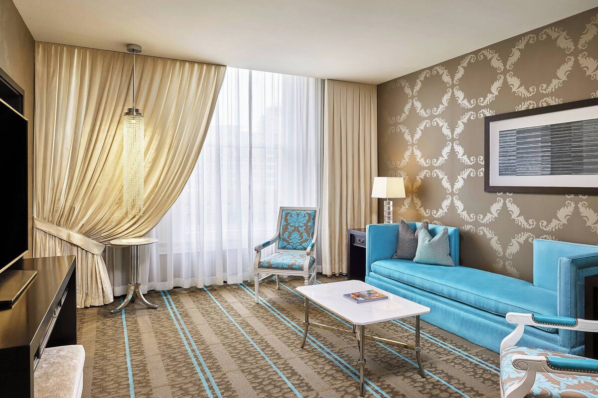 A hotel room with a blue sofa, a white coffee table, a blue armchair, a TV, and a large window with curtains.