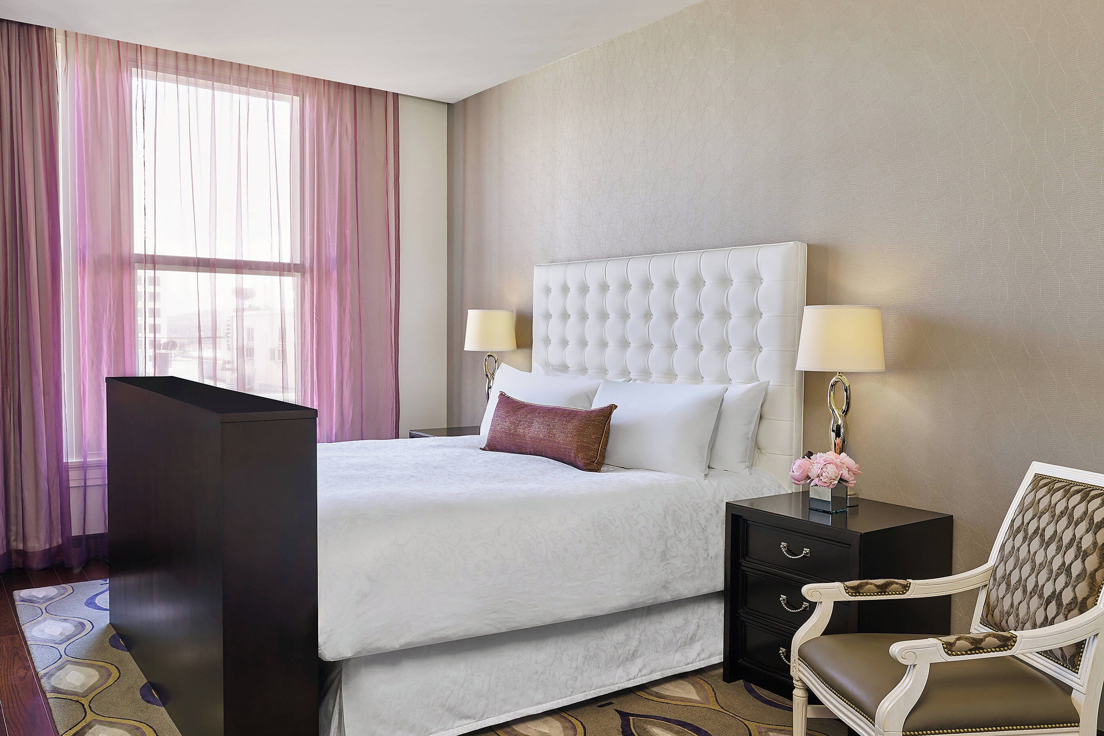 Une chambre d’hôtel avec un grand lit, deux tables de chevet, une chaise et une fenêtre avec des rideaux légers.