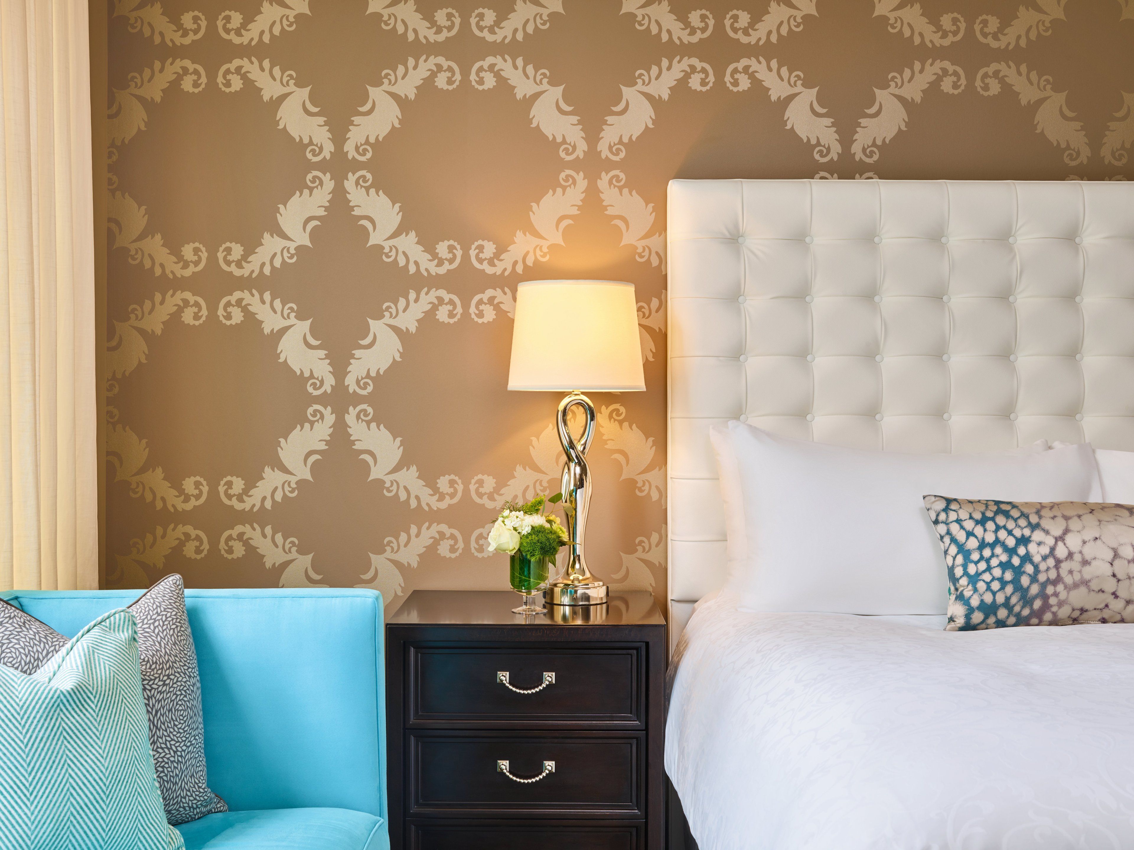 A hotel room with a tufted headboard, a bedside lamp, a floral vase, a blue sofa, and a patterned pillow.