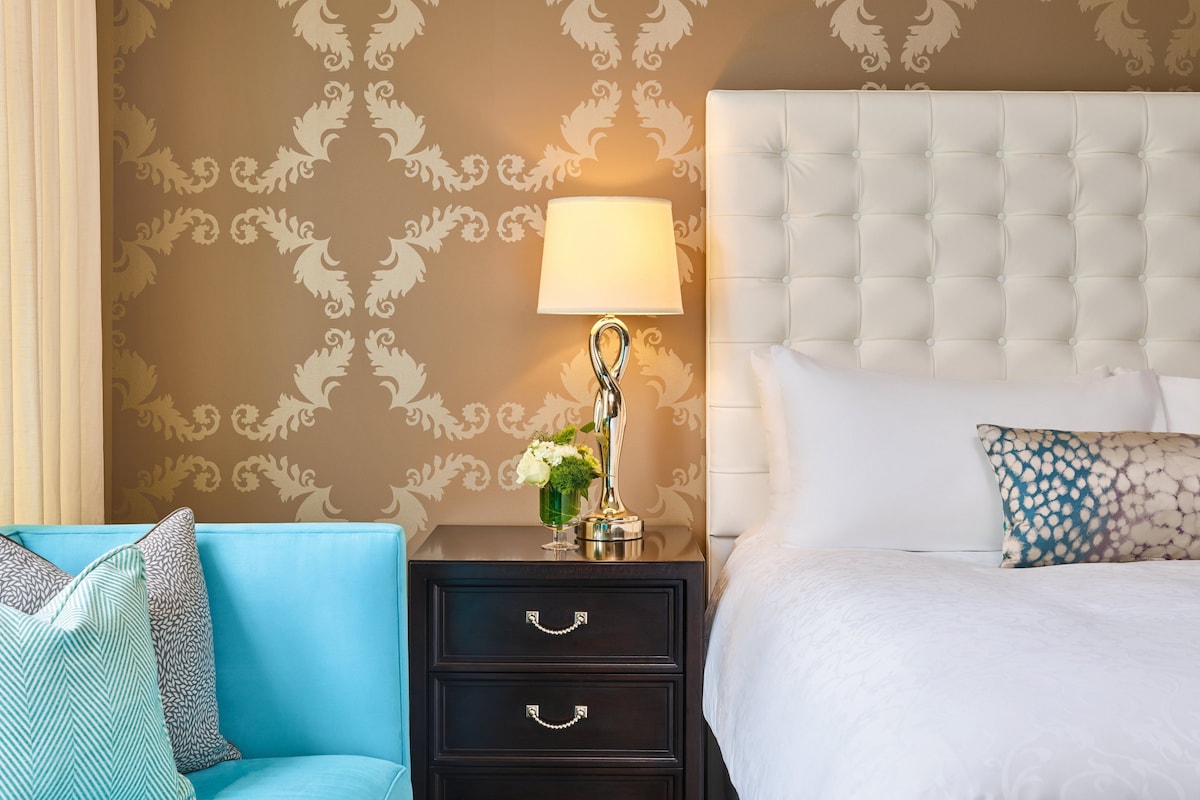 A hotel room with a tufted headboard, a bedside lamp, a floral vase, a blue sofa, and a patterned pillow.