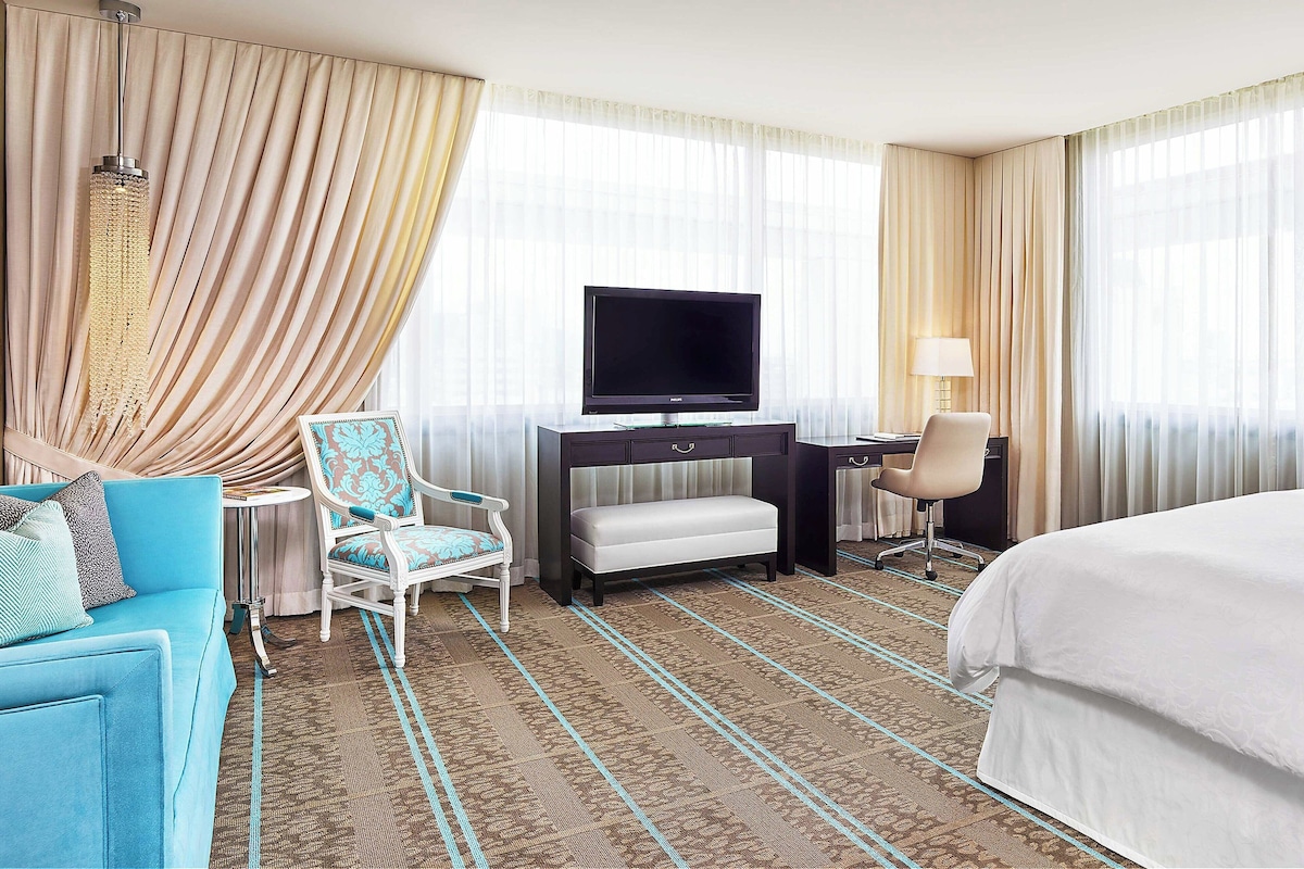 A hotel room with a large bed, a desk with a flat-screen TV, a blue sofa, two chairs, and a patterned carpet.