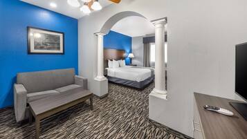 A hotel room with a blue accent wall, a bed, a sofa, a desk with a TV, and a ceiling fan.