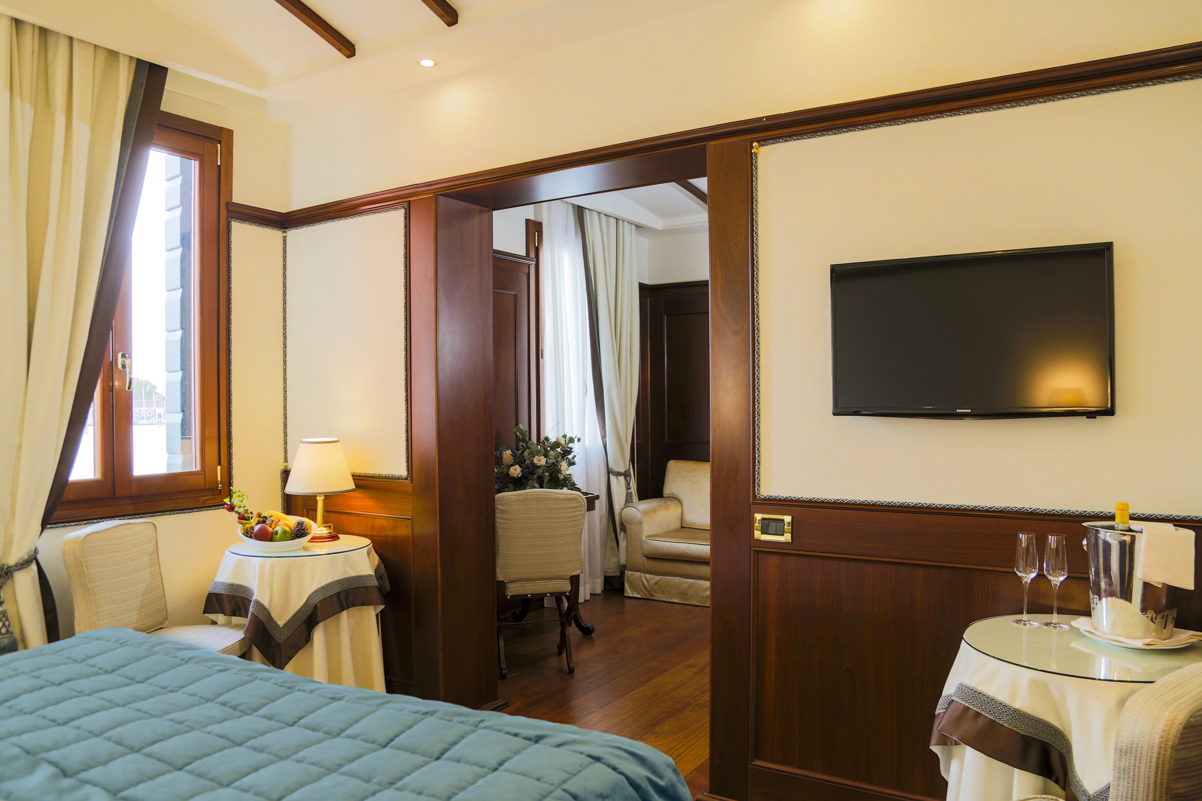 Habitación de hotel con cama, escritorio con lámpara, silla, televisor y pequeña mesa con vasos y botella.