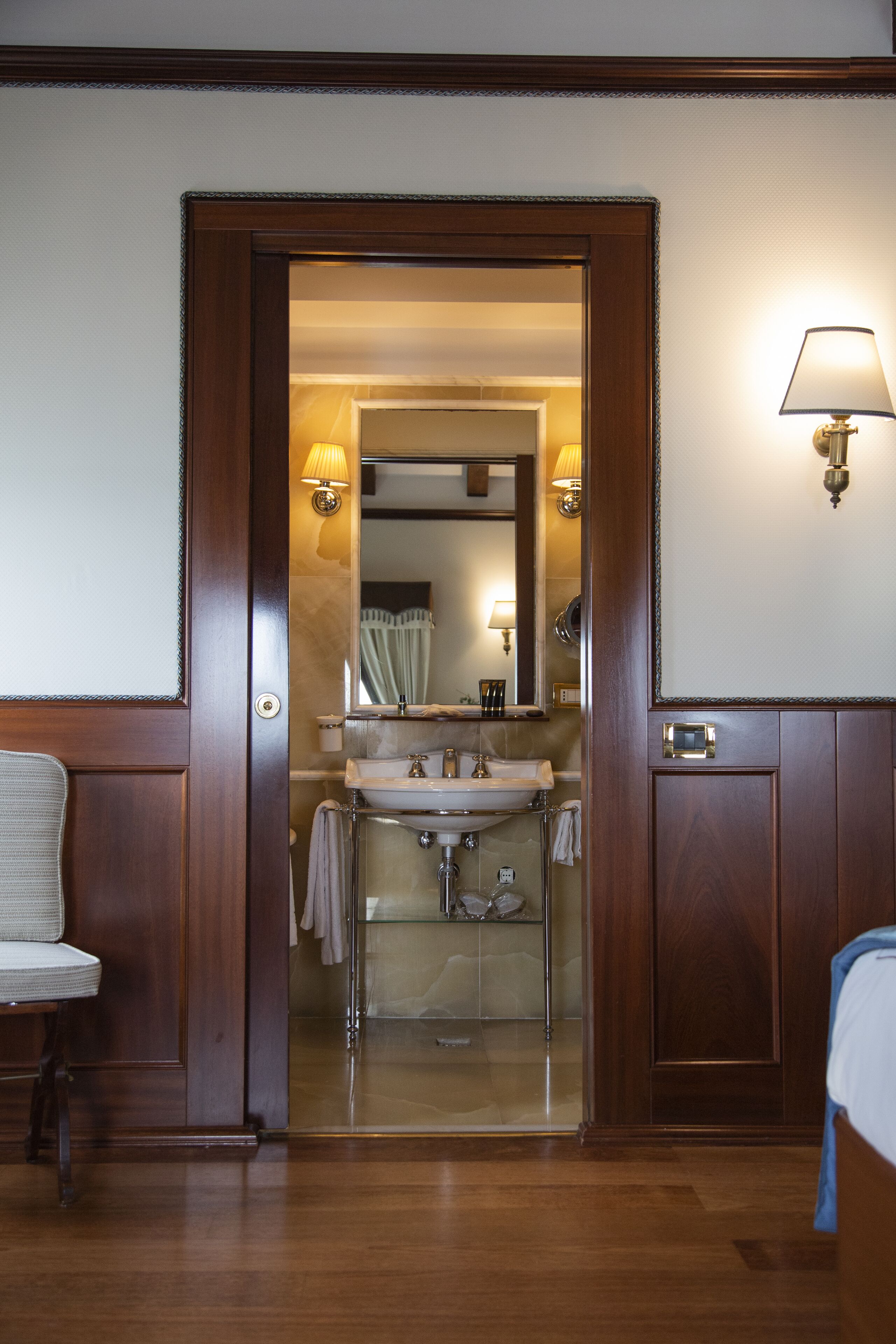 A hotel room with a wooden door leading to a bathroom. The bathroom has a sink, mirror, and towel rack. There is a bed with a blue blanket and a chair with a white cushion.
