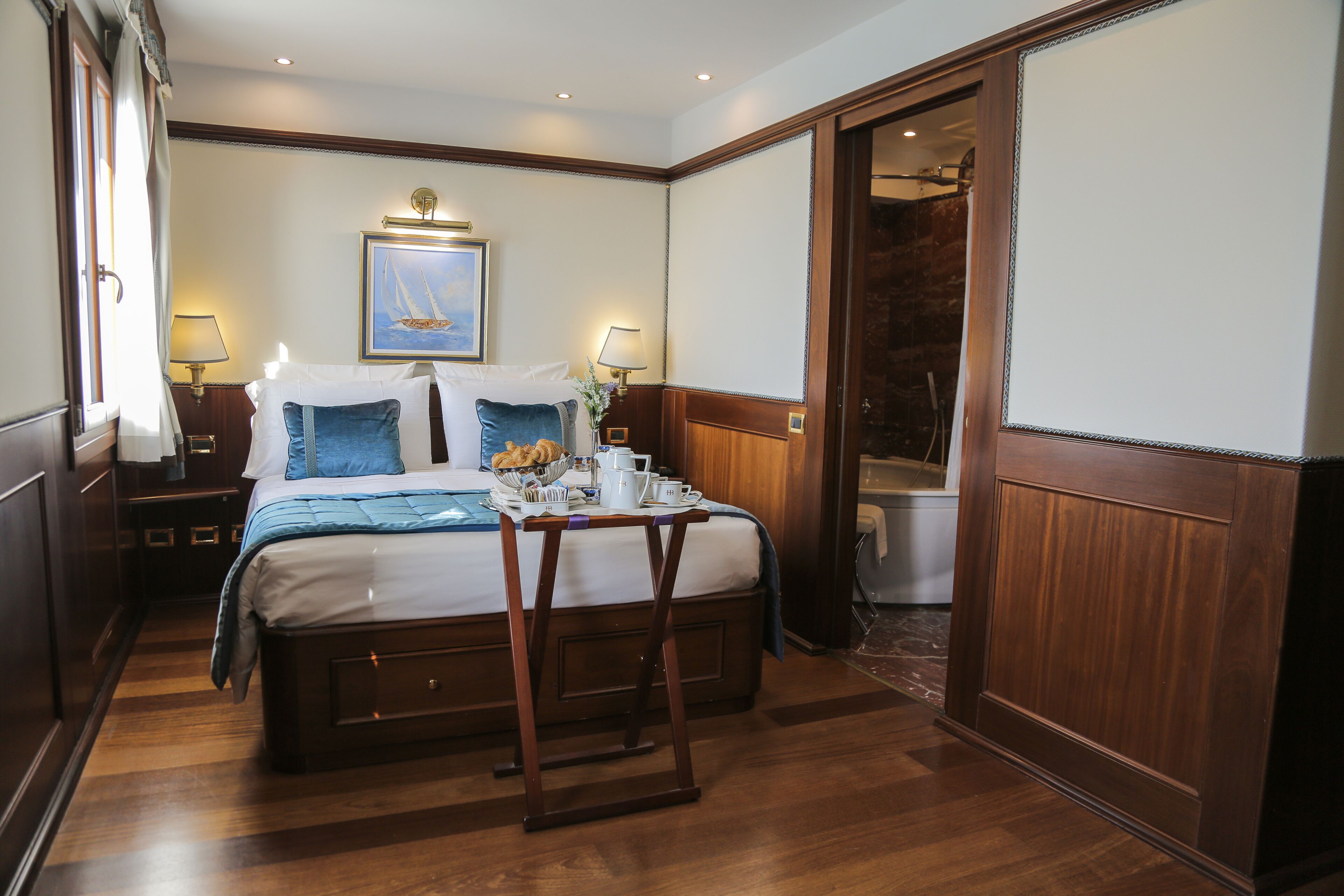 Une chambre d’hôtel avec un lit en bois, une petite table sur laquelle sont disposés des articles pour le petit-déjeuner, un tableau représentant un voilier, et une salle de bain visible à travers une porte ouverte.