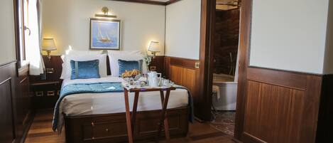 Une chambre d’hôtel avec un lit en bois, une petite table sur laquelle sont disposés des articles pour le petit-déjeuner, un tableau représentant un voilier, et une salle de bain visible à travers une porte ouverte.
