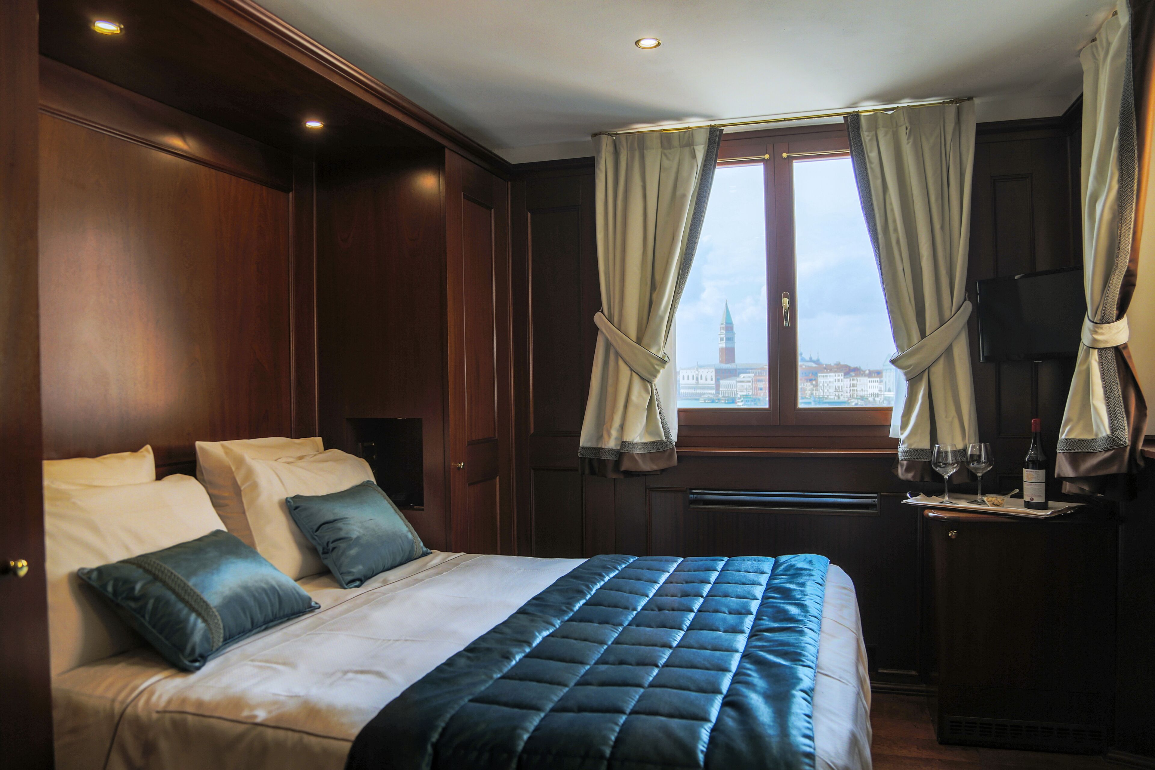 Habitación de hotel con una cama grande, una mesita con una botella y vasos, y una ventana con cortinas.