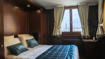 Habitación de hotel con una cama grande, una mesita con una botella y vasos, y una ventana con cortinas.