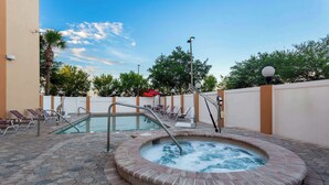 Ein Whirlpool, umgeben von Liegestühlen und einem Pool, mit blauem Himmel und Bäumen im Hintergrund.