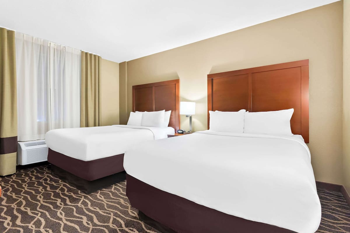 Ein Hotelzimmer mit zwei Betten, einem großen und einem kleineren, beide mit weißen Bettwäsche. Ein Fenster mit beigen Vorhängen, eine Wand-Klimaanlage und ein Teppich mit Muster.