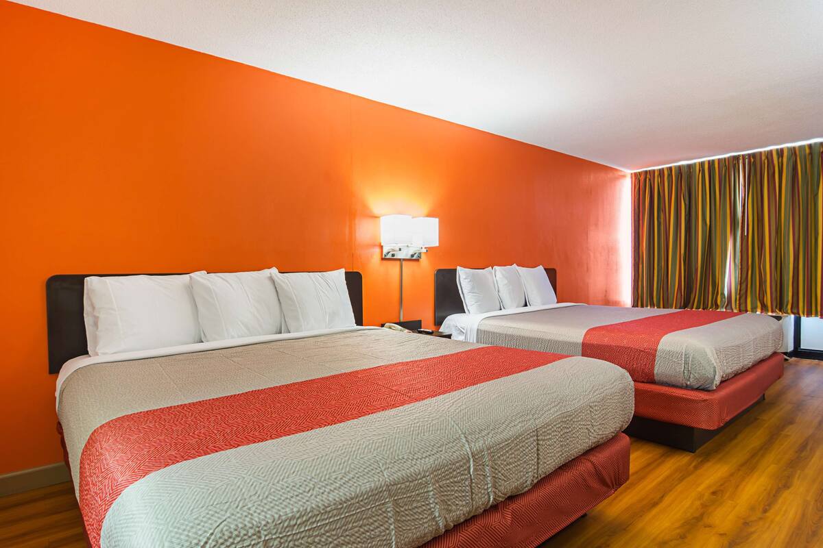 A hotel room with two beds, a nightstand, a lamp, and a window with curtains.