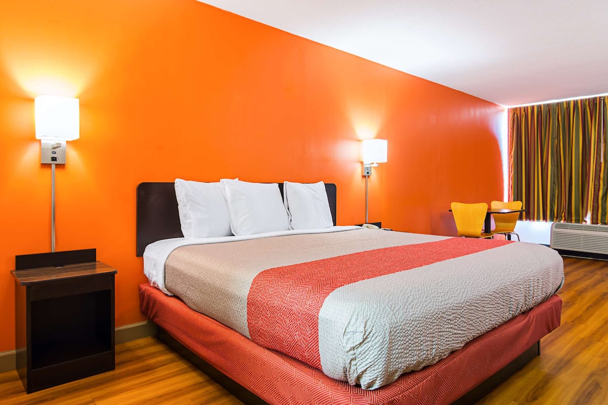 A hotel room with a large bed, orange walls, two bedside lamps, a small table, and a chair.