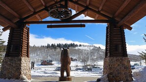 Ein hölzerner Pavillon mit Steinunterbau, einer Bärenstatue und Blick auf eine verschneite Berglandschaft.
