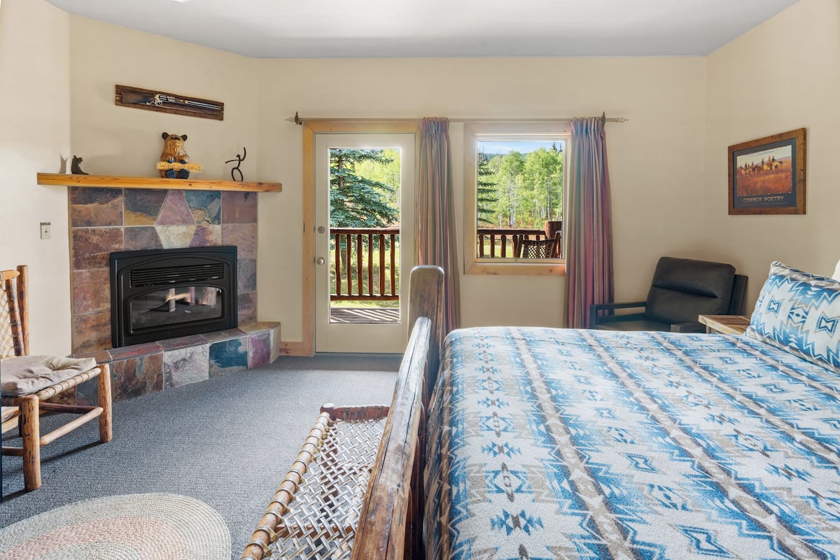 A bedroom with a fireplace, a bed with patterned bedding, a chair, and a view of the outdoors.