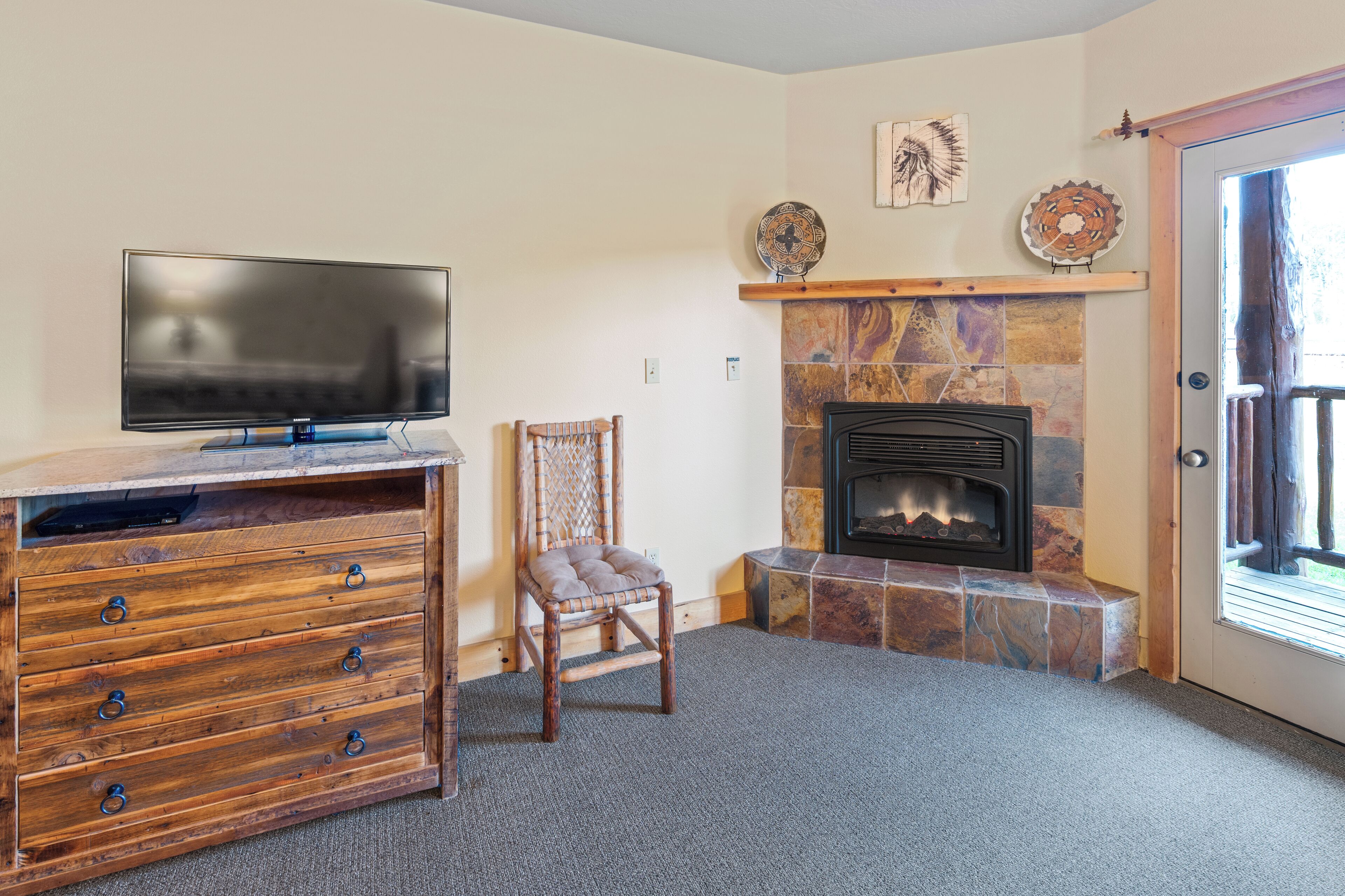A living room with a flat-screen TV, wooden entertainment center, stone fireplace, and a door leading outside.