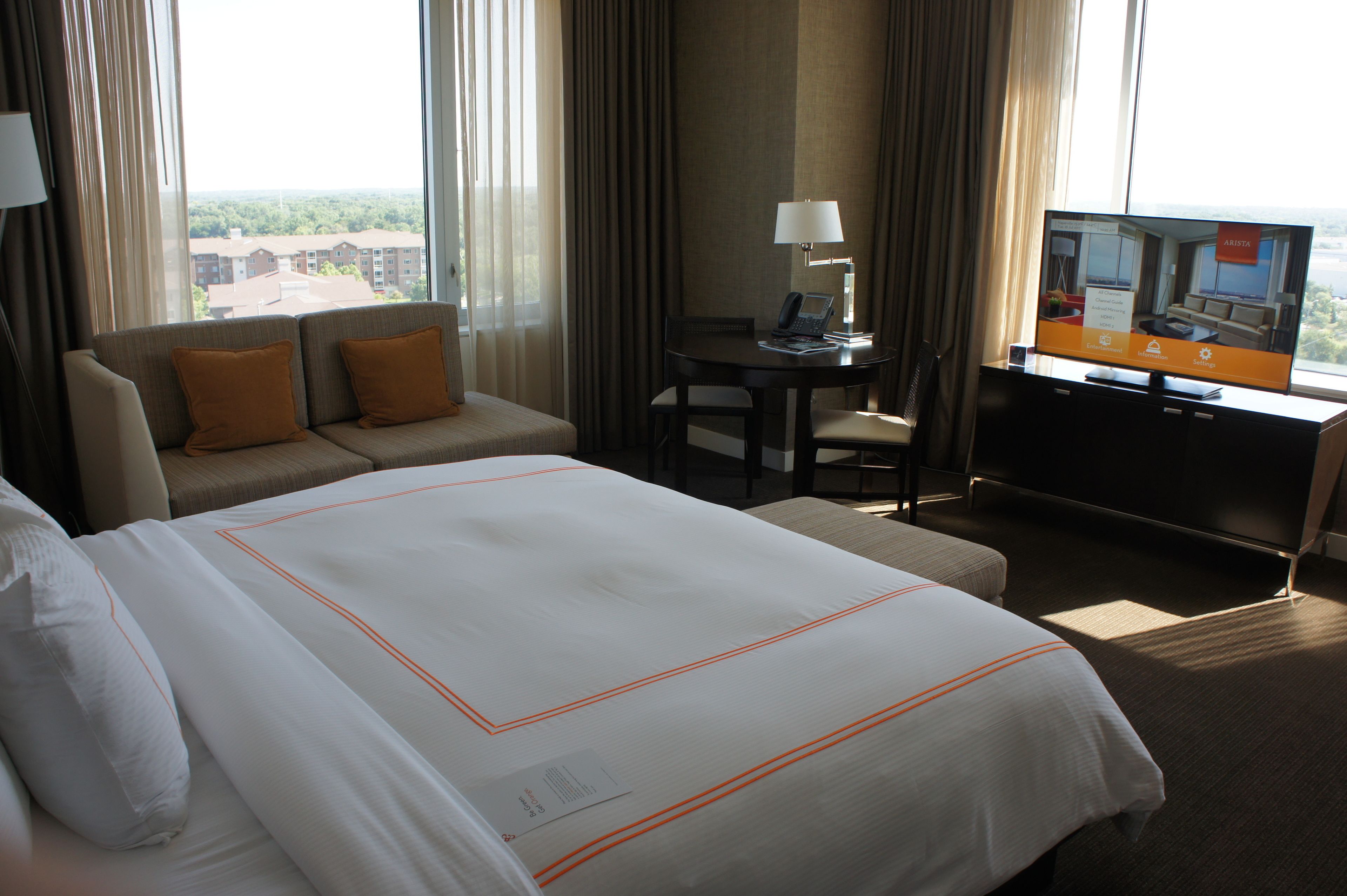 A hotel room with a large bed, a sofa, a desk with a lamp, and a TV displaying room information.