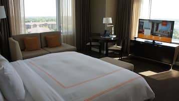 A hotel room with a large bed, a sofa, a desk with a lamp, and a TV displaying room information.