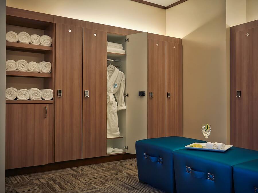 A hotel room with a large wardrobe, a seating area with a table, and neatly rolled towels on shelves.