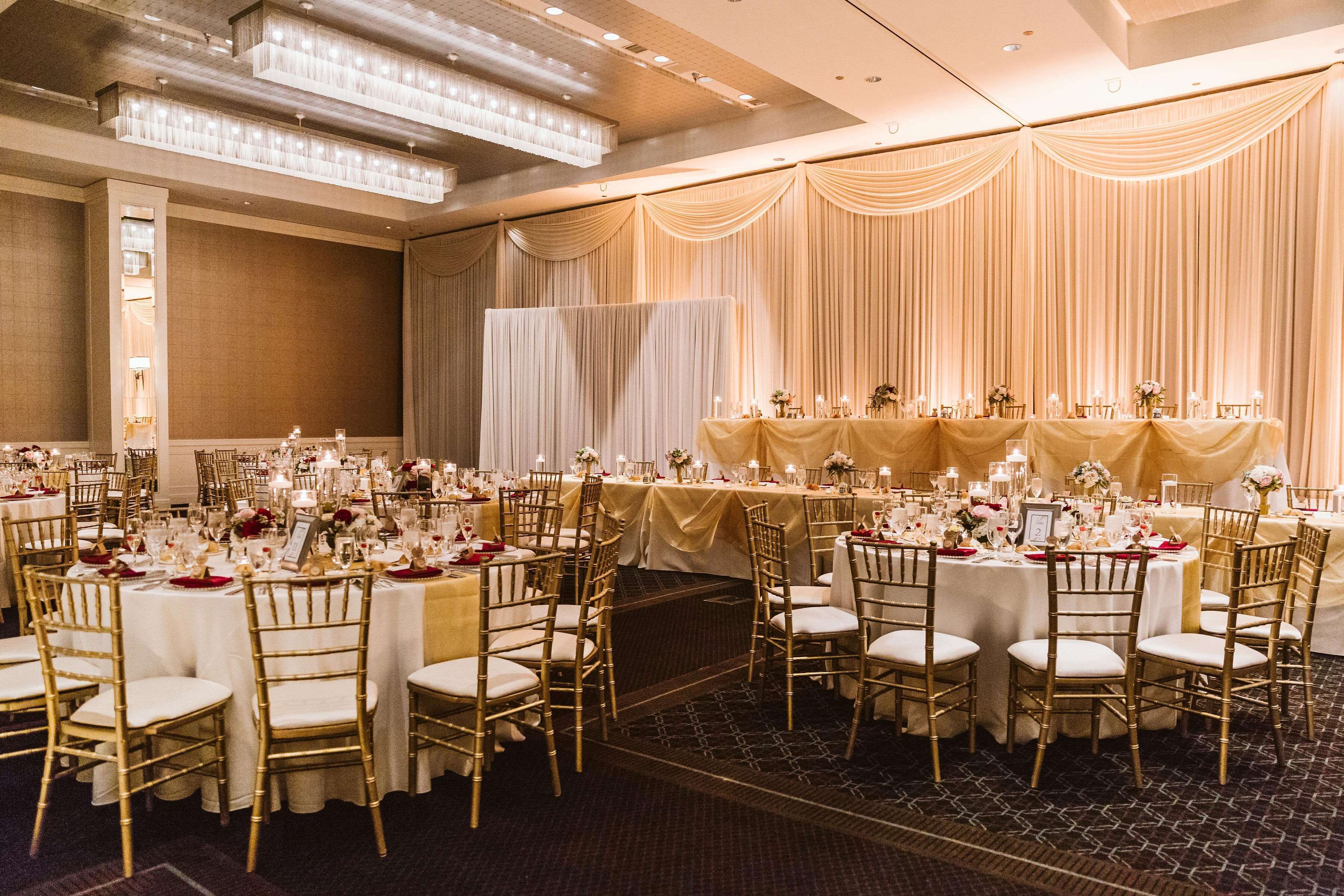Un salón de banquetes con mesas redondas cubiertas con manteles blancos, sillas doradas y centros de mesa florales.