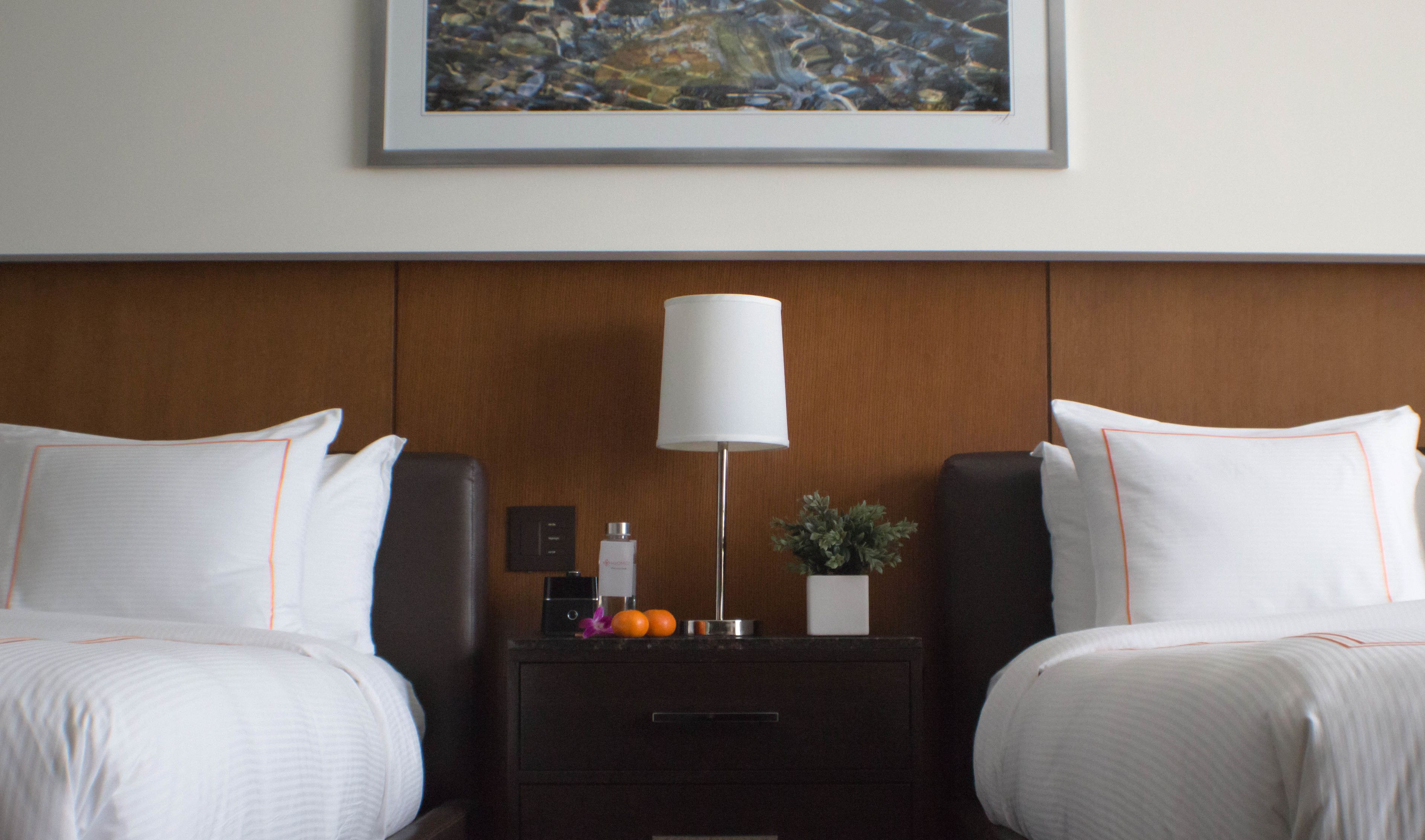 A hotel room with two beds, a nightstand with a lamp, a small plant, and a framed artwork on the wall.