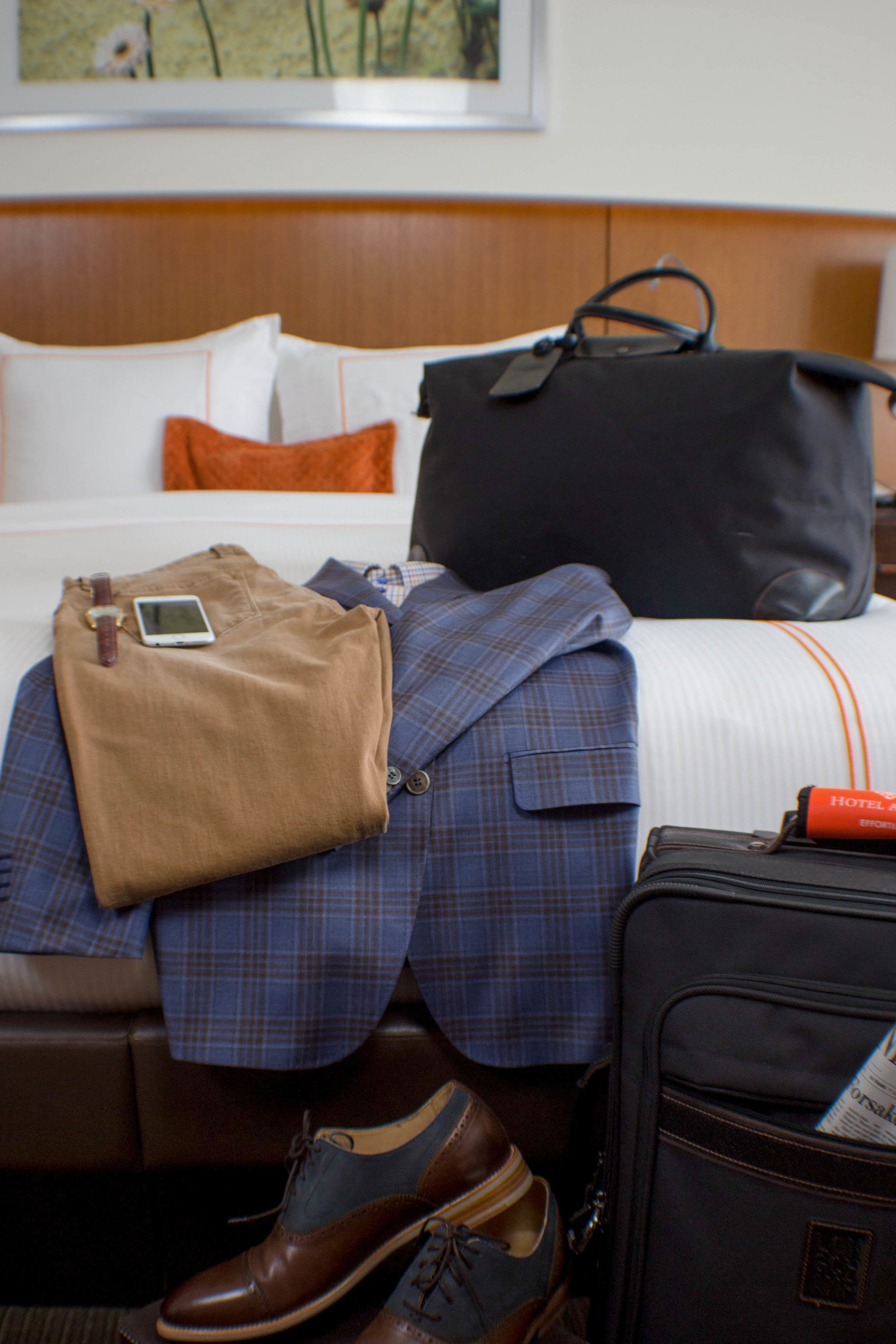 A neatly made bed with a pillow, a black bag, a brown bag, a smartphone, a pair of shoes, and a suitcase.