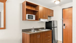 A compact kitchen with wooden cabinets, a microwave, a stainless steel refrigerator, and a sink. - Welcome Suites (O'Fallon)