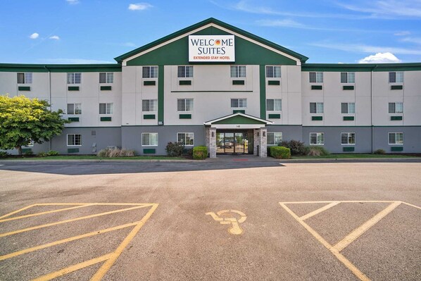 A hotel with a green and white facade, a sign reading "Welcome Suites Extended Stay Hotel," and a parking lot with marked handicapped spaces. - Welcome Suites (O'Fallon)