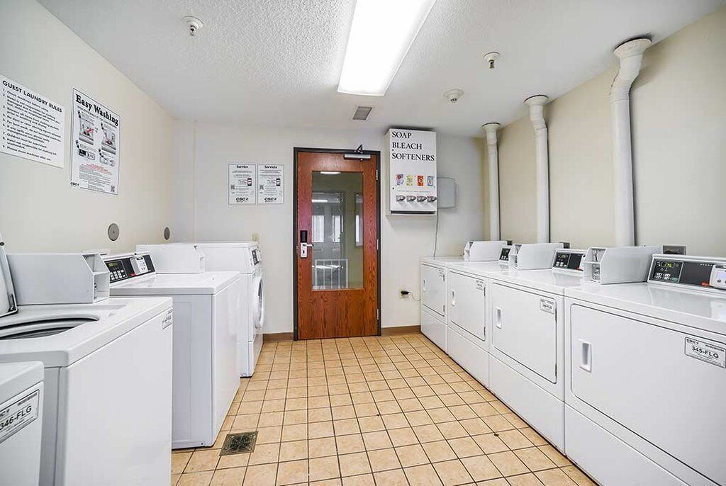 Une buanderie équipée de plusieurs machines à laver et de plusieurs sèche-linge, avec un sol carrelé et des affiches d’information accrochées aux murs.