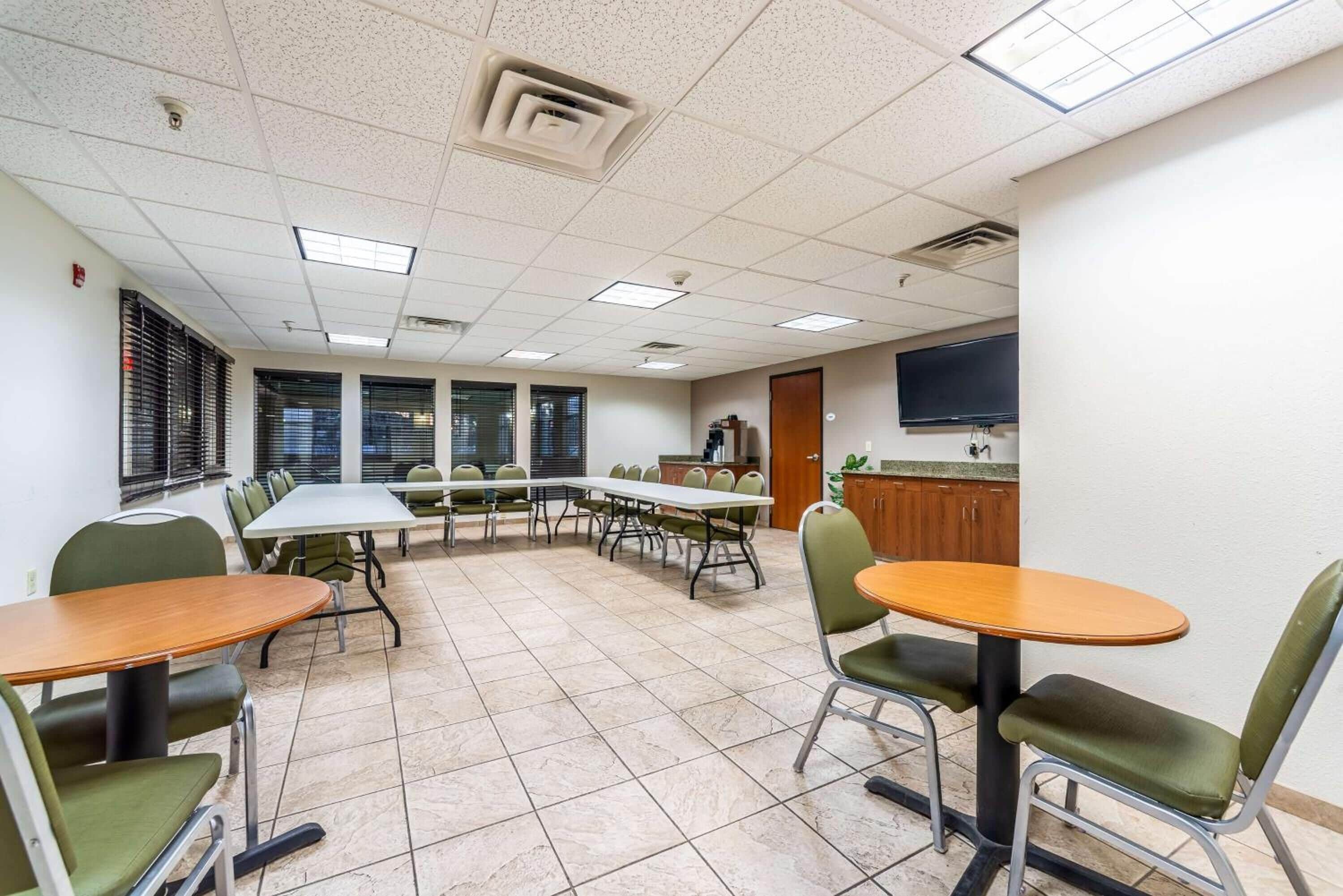A conference room with multiple tables and chairs, a television on the wall, and a kitchenette area.