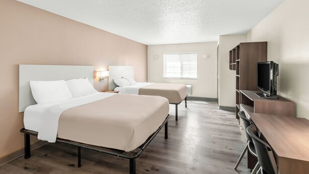 A hotel room with two beds, a desk, a TV, and a window with blinds. - Welcome Suites (O'Fallon)