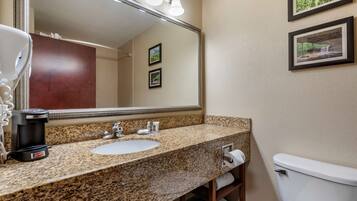 A bathroom with a granite countertop sink, a coffee maker, a mirror with lights, and framed pictures on the wall.