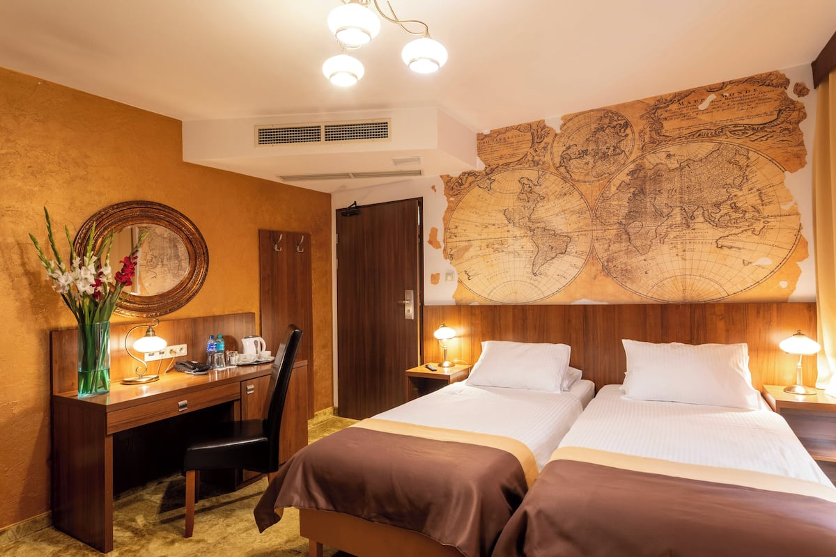 A hotel room with a wooden desk, two beds, a large world map on the wall, a mirror, and a vase with flowers.