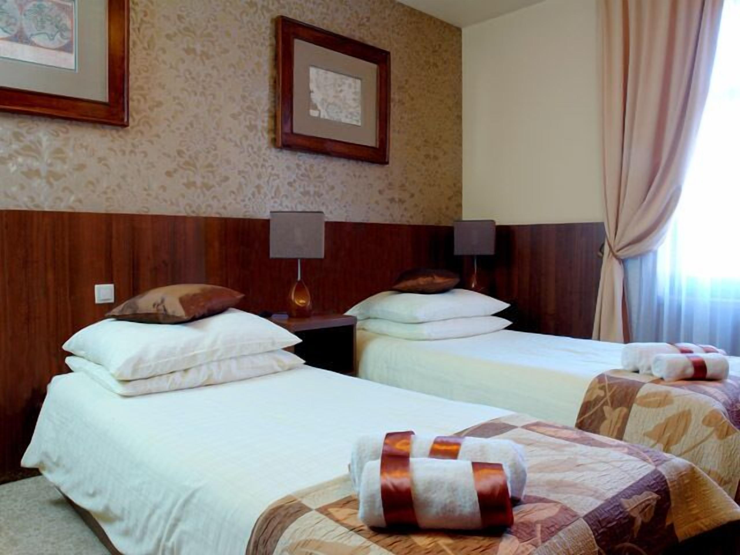 Habitación de hotel con dos camas, cada una con ropa blanca y almohadas, cabeceros de madera, un cuadro en la pared y una lámpara en la mesita de noche.