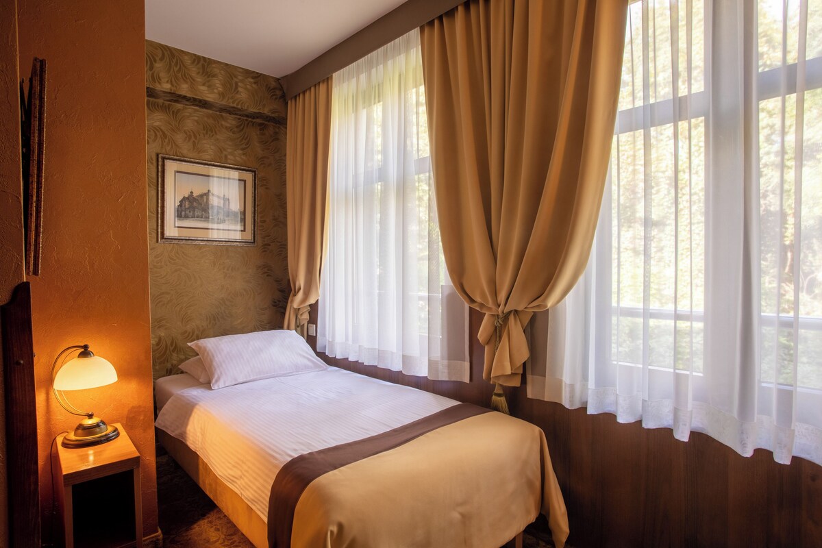 A hotel room with a bed, a nightstand with a lamp, a framed picture on the wall, and large windows with curtains.