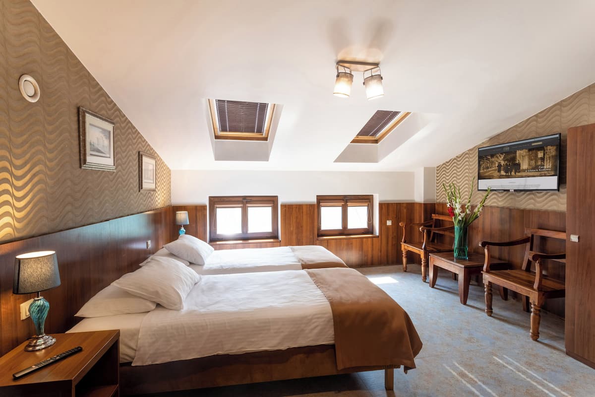 A hotel room with two beds, a TV, a desk with a lamp, and a wooden bench.